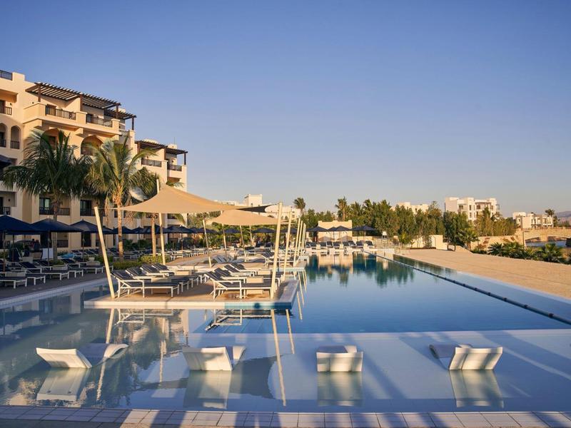 Großer Infinity-Pool mit Liegestühlen und Palmen vor einer hellen Hotelanlage bei klarem Himmel.