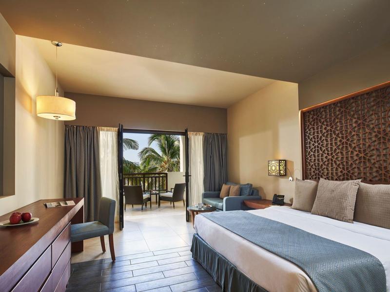 Helles Hotelzimmer mit Doppelbett, Holzkommode, Sitzbereich und Balkon mit Palmenblick.