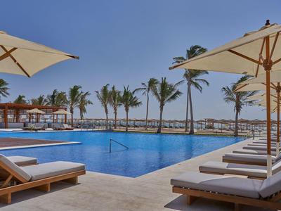 Area piscina moderna con lettini e ombrelloni, palme e mare sullo sfondo.