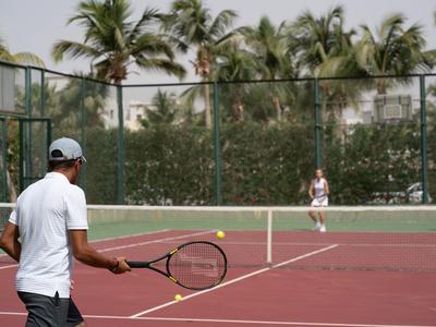 Twee mensen spelen tennis op een buitenbaan met palmbomen op de achtergrond.