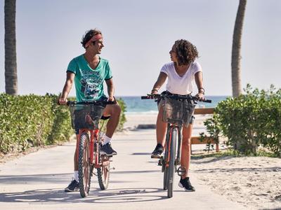 Twee personen fietsen op een zanderig pad bij het strand onder een heldere lucht.