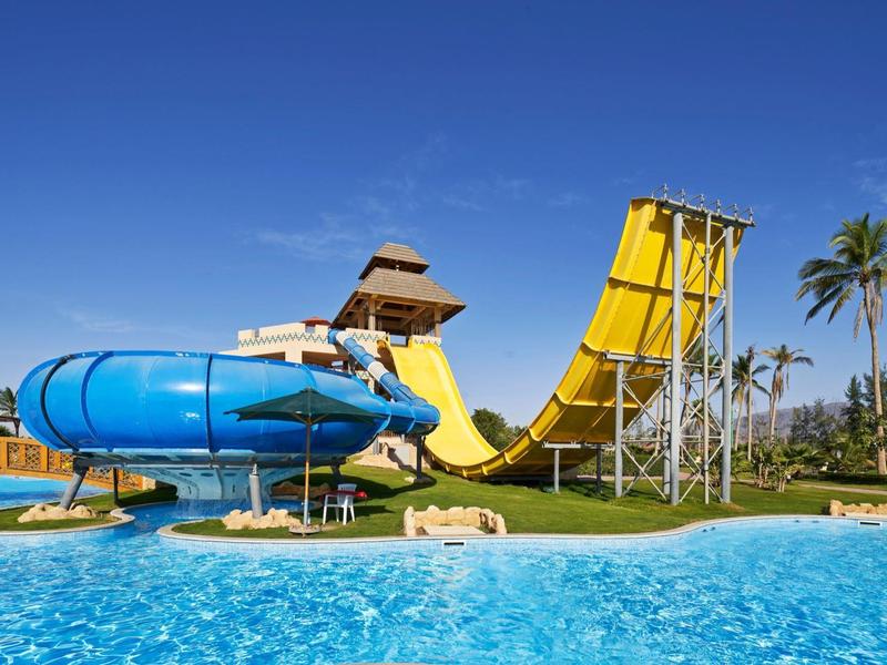 Parco acquatico con scivoli d'acqua blu e gialli accanto a una piscina sotto un cielo limpido.