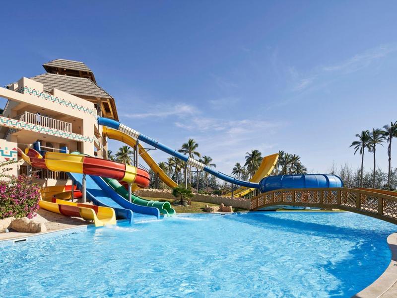 Parco acquatico con scivoli colorati, grande piscina e palme sotto un cielo sereno.