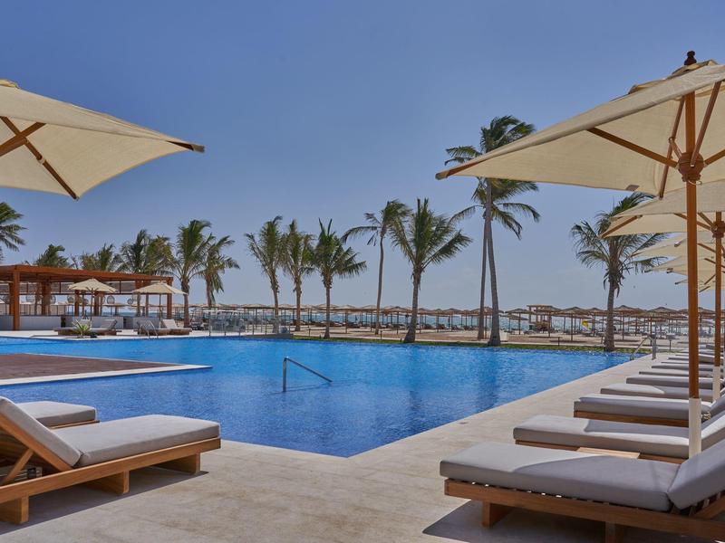 Area piscina moderna con lettini e ombrelloni, palme e mare sullo sfondo.
