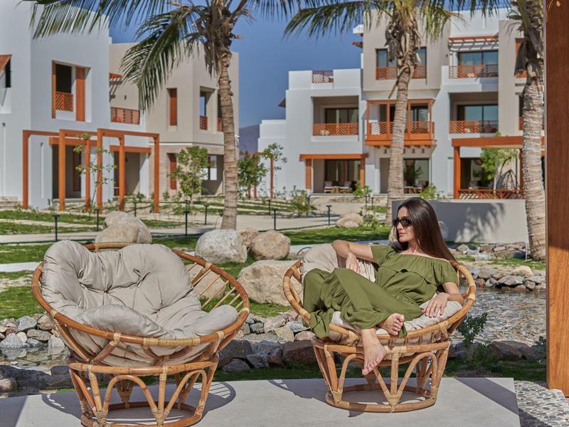 Una donna è seduta rilassata su una sedia di vimini su una terrazza con vista su un giardino e edifici alberghieri moderni.