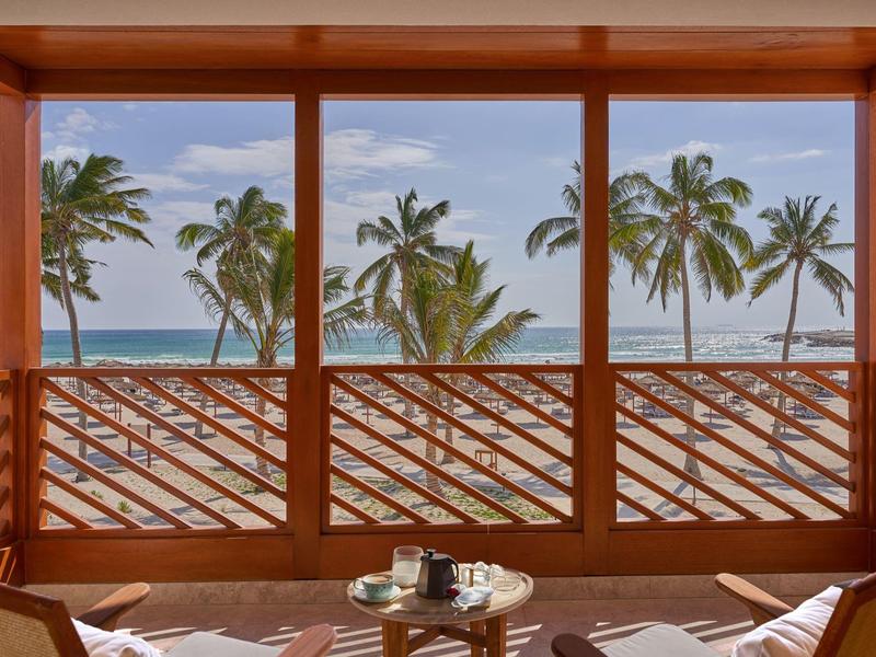 Vista di palme e mare attraverso un balcone vetrato con grata in legno.