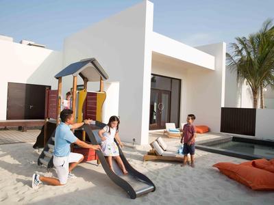 Family playing on a sandy playground next to a modern house with a pool.