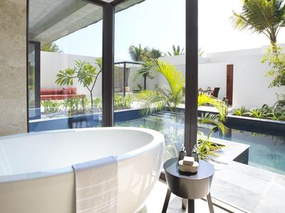 Freestanding bathtub with towel next to glass wall overlooking tropical pool and garden.