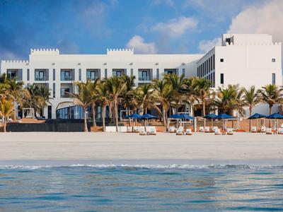 White beach hotel with palm trees and lounge chairs under blue sky.