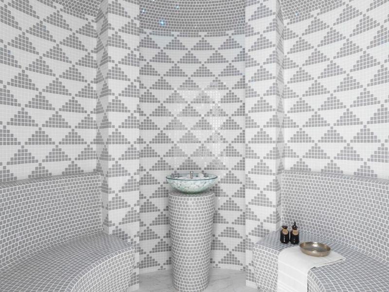 Modern steam room with gray mosaic tile walls, curved benches, and central pedestal sink.
