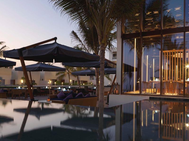 Modern hotel pool area at dusk with palm trees and illuminated glass walls.