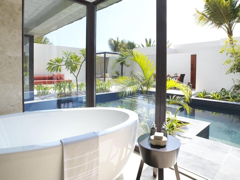 Freestanding bathtub with towel next to glass wall overlooking tropical pool and garden.