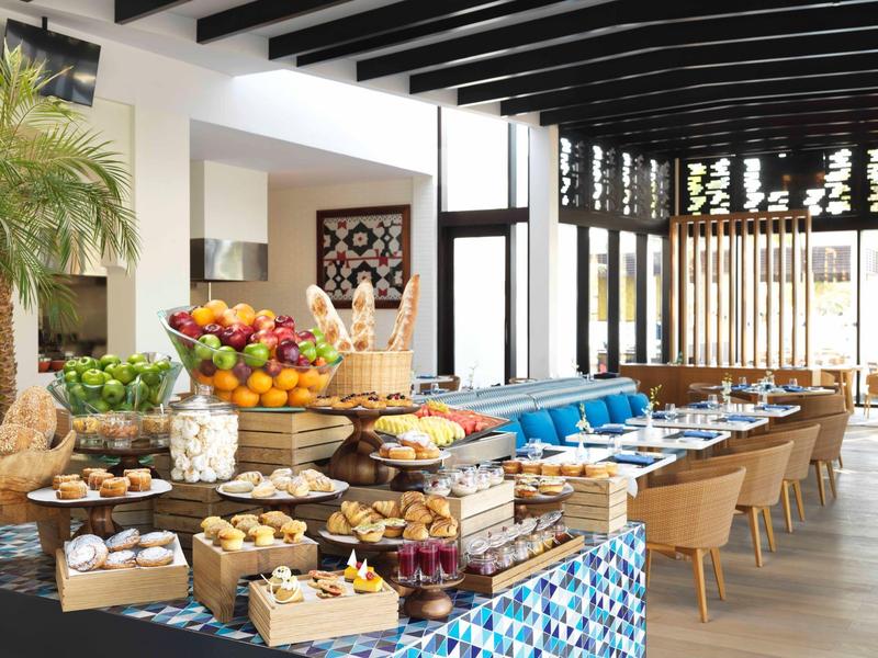 Modern restaurant with wooden furniture, fresh fruit, and set tables in a bright space.