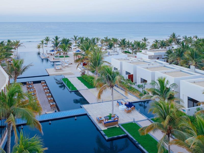 Modern beachfront resort with pools, palm trees, and white villas by the calm sea.