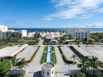 Hotelanlage mit weißem Gebäude, großen Parkplätzen, Palmen und blauem Himmel am Meer im Hintergrund.