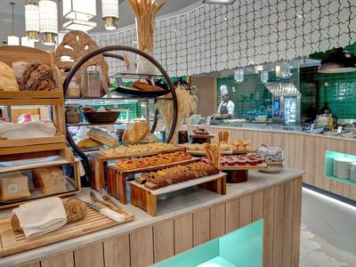 Modernes Café mit hellem Holztresen, frischem Gebäck und grüner Fliesenwand im Hintergrund.