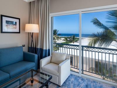 Helles Wohnzimmer mit blauer Couch, weißem Sessel und großem Fenster mit Blick auf Palmen und Meer.