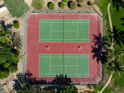 Luchtfoto van een buitentennisbaan met groene speeloppervlakte en rode omgeving, omringd door bomen.