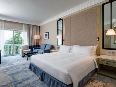 Lichte hotelkamer met groot bed, bank, tapijt en balkon met groen uitzicht.