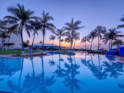 Ein klarer Pool spiegelt Palmen und den bunten Abendhimmel an einer tropischen Küste.