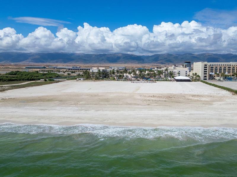 Weitläufiger Sandstrand mit leichtem Wellengang, dahinter Stadt und Berge unter blauem Himmel.