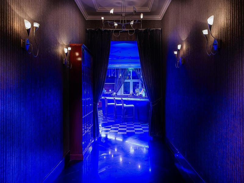 Flur mit dunklen Wänden, blauer Beleuchtung und Blick auf einen hell erleuchteten Raum dahinter.