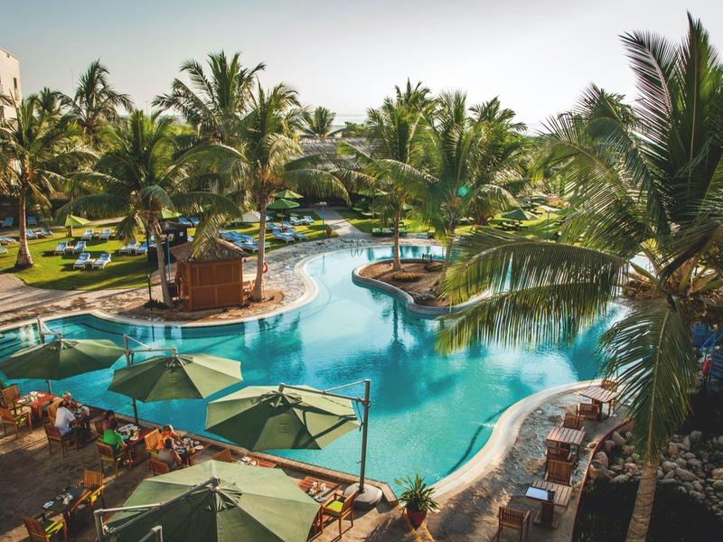 Großer Pool mit blauem Wasser, umgeben von Palmen, Liegestühlen und Sonnenschirmen in tropischer Anlage.