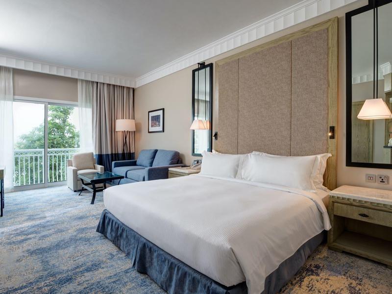 Helles Hotelzimmer mit großem Bett, Sofa, Teppichboden und Fenster mit Blick ins Grüne.