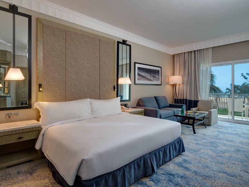 Modernes Hotelzimmer mit großem Bett, Sofa, Teppichboden und Balkon mit Blick ins Grüne.