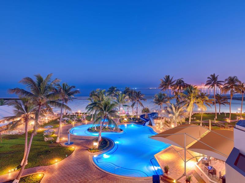 Luxuriöser Poolbereich mit Palmen, beleuchtet bei Dämmerung, neben ruhigem Meer und blauem Himmel.