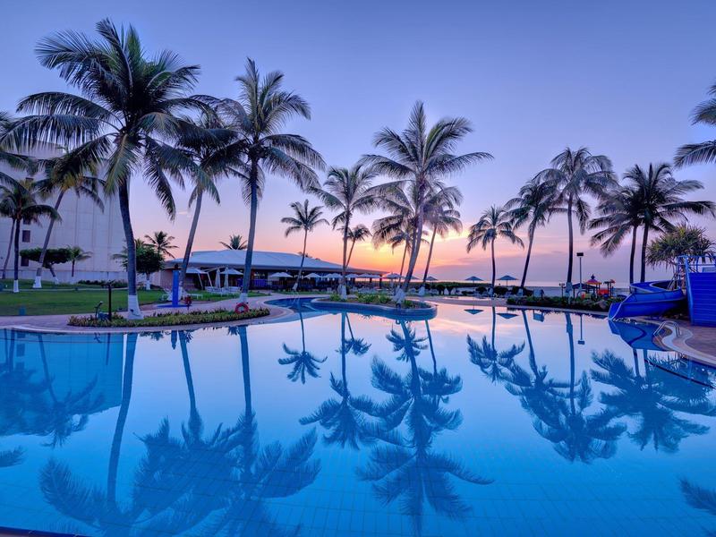 Ein klarer Pool spiegelt Palmen und den bunten Abendhimmel an einer tropischen Küste.