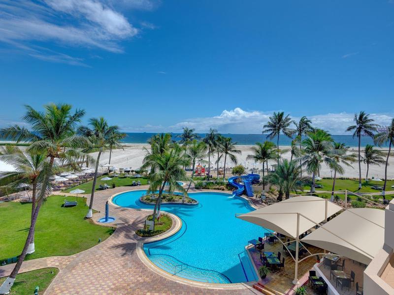 Hotelpool mit Palmen, Sonnenschirmen und Blick auf weißen Sandstrand und blauen Himmel.