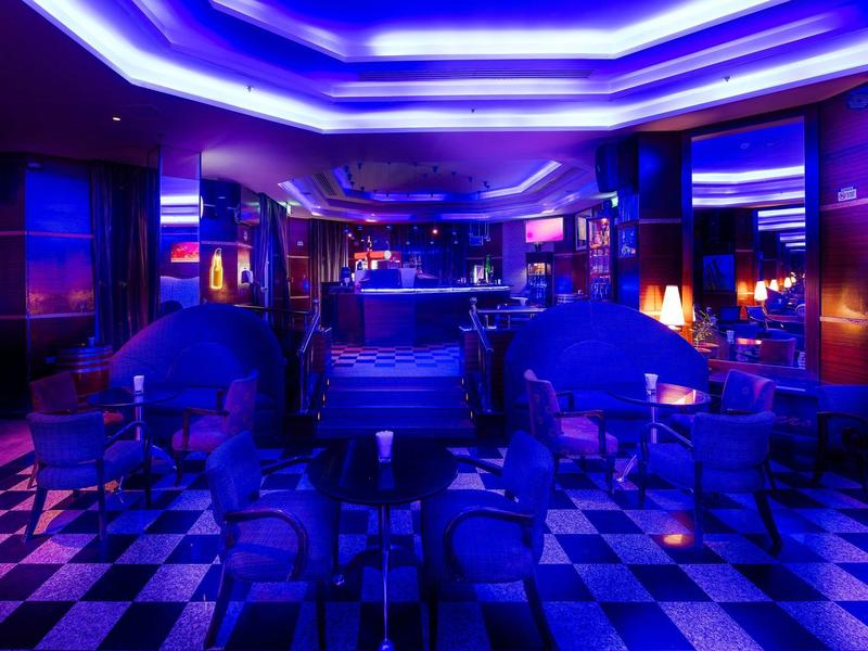 Dunkle Bar mit blauer Neonbeleuchtung, Sitzecken und schwarz-weißem Schachbrettboden.