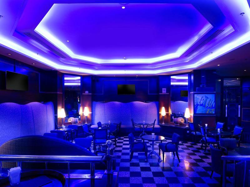 Dunkler Clubraum mit blauer Beleuchtung, Sitzgruppen, kleinem Flügel und Schachbrettboden.