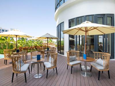 Terrasse mit Holzstühlen, runden Tischen und großen Sonnenschirmen vor modernem Gebäude.