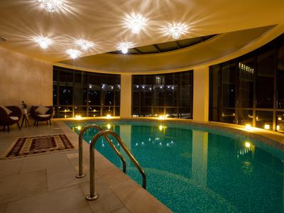 Innenraum mit rundem Schwimmbecken, warmem Licht, großen Fenstern und Sitzbereich mit Stühlen und Teppich.