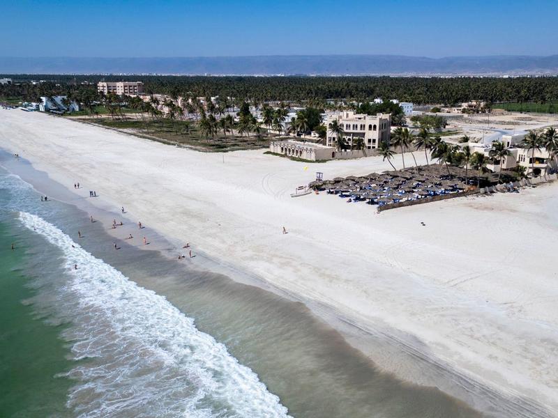 Weißer Sandstrand mit Palmen, Wellen im blauen Meer und Gebäuden im Hintergrund.