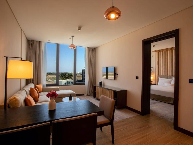 Moderner Wohnbereich mit Sofa, Tisch, Stühlen, Lampen und Blick auf ein Schlafzimmer, große Fenster.