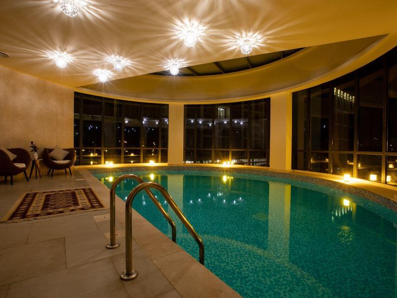 Innenraum mit rundem Schwimmbecken, warmem Licht, großen Fenstern und Sitzbereich mit Stühlen und Teppich.