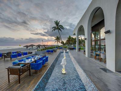 Terrasse am Meer mit blauen Sitzkissen, Palmen und Wasserlauf neben weißem Gebäude mit Bögen.