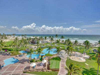 Vista di un'area piscina tropicale con palme e spiaggia sotto un cielo azzurro.