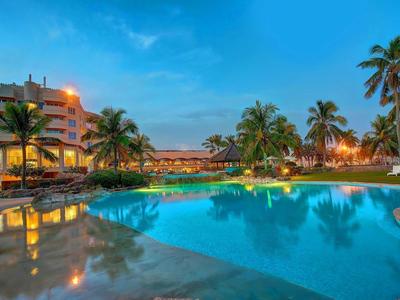 Vista serale di un hotel con piscina illuminata e palme sotto un cielo limpido.