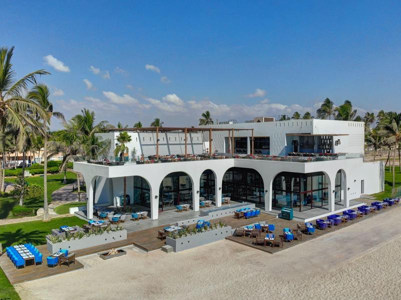 Hotel moderno bianco sulla spiaggia con piscina, lettini e palme sotto un cielo azzurro.