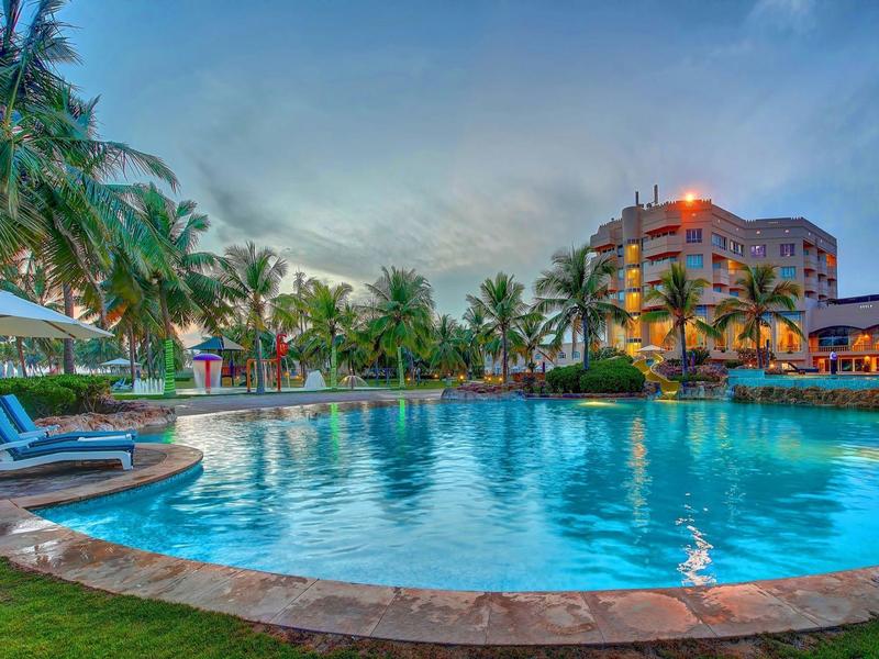 Piscina rotonda dell'hotel circondata da palme, lettini e edificio pluripiano al tramonto.