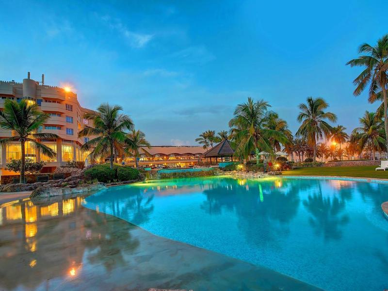 Vista serale di un hotel con piscina illuminata e palme sotto un cielo limpido.