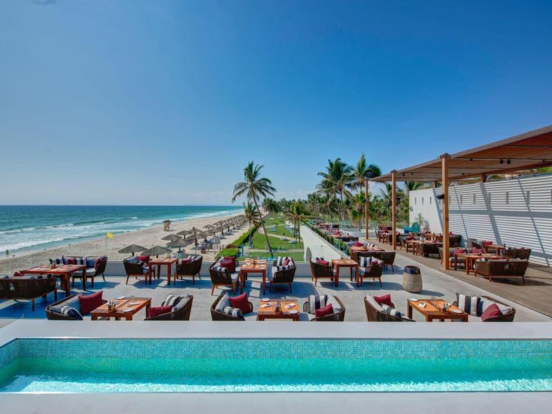Resort sulla spiaggia con piscina, aree lounge, lettini e palme sotto un cielo azzurro limpido