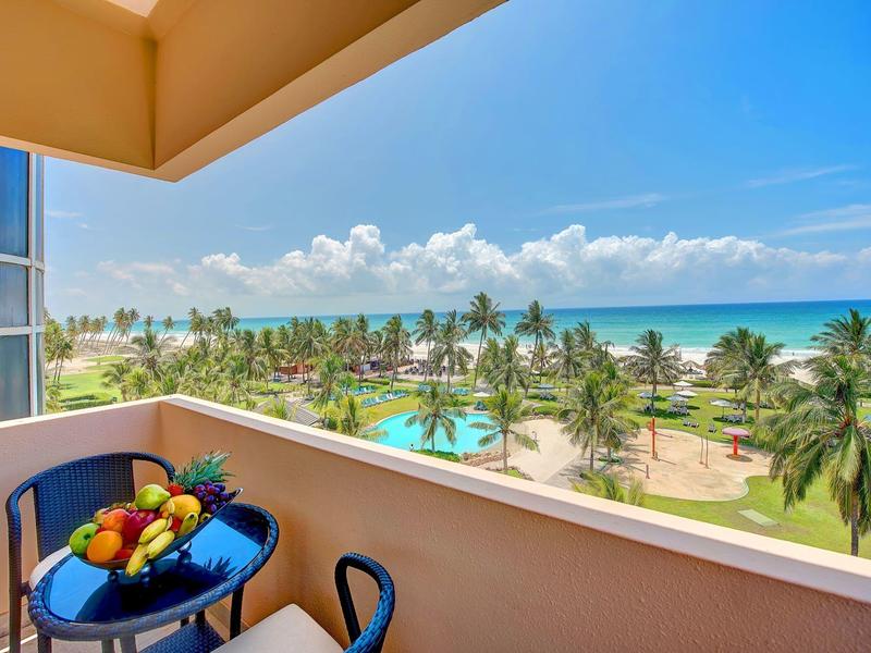 Balcone con tavolo e frutta, vista su palme, spiaggia e mare sotto un cielo limpido.