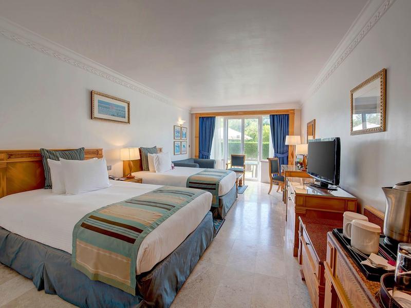 Camera d'hotel luminosa con due letti, TV, tavolo e balcone con vista sul giardino.