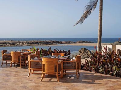Strandrestaurant mit Holztischen und -stühlen vor Meer und Wellenbrecher bei klarem Himmel.