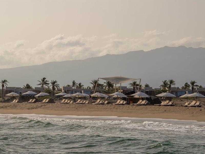Plage avec chaises longues et parasols devant des montagnes au bord d'une mer calme.